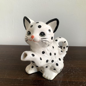 Puede incluir: Una tetera de cerámica blanca con forma de gato manchado con manchas negras. La tetera tiene un pico en forma de pata de gato.