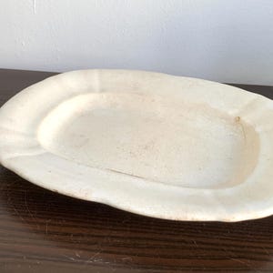 May include: A white ceramic oval serving dish with a slightly raised rim. The dish has a smooth, slightly textured surface.