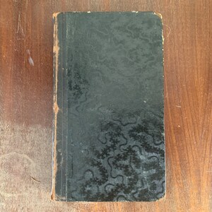 Pode incluir: Um livro antigo, pequeno e retangular, com uma capa preta com um padrão texturizado e ondulado. As bordas do livro mostram sinais de desgaste e envelhecimento. O livro está sobre uma superfície de madeira.