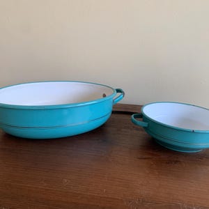 Könnte beinhalten: Zwei Vintage-Emaille-Schalen. Die Schalen sind oval geformt mit türkisfarbenem Außen und weißem Innenraum. Jede Schale hat zwei Griffe und ein dünnes schwarzes Streifendetail. Die Schalen werden auf einer Holzoberfläche präsentiert.