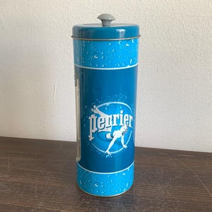 Può includere: Un barattolo Perrier alto e cilindrico con un design blu e bianco. Il barattolo presenta il logo Perrier e una grafica di una donna che si tuffa. Il coperchio e il bordo superiore sono turchese e il corpo è blu scuro.