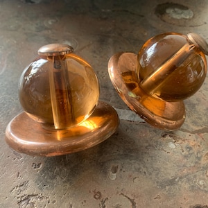 Könnte beinhalten: Zwei bernsteinfarbene Glasknöpfe mit Kupfersockeln. Die Knöpfe sind rund und haben eine glatte, glänzende Oberfläche. Die Sockel sind rund und haben eine leicht strukturierte Oberfläche.
