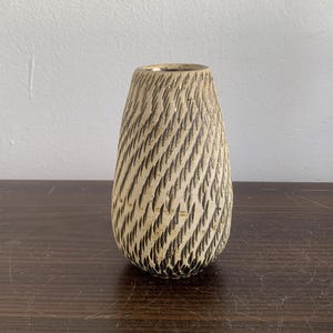 Op de afbeelding: Een kleine, crèmekleurige keramische vaas met een getextureerd, zwart-wit patroon. De vaas heeft een afgeronde, licht uitlopende basis en een smalle opening.
