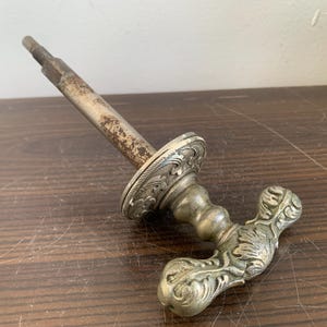 May include: A silver-toned door handle with an ornate design. The handle has a long, thin shaft and a decorative knob with a floral pattern.