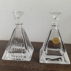 Pode incluir: Duas garrafas de perfume de vidro transparente com corpos em forma de pirâmide e rolhas geométricas. Uma garrafa tem um rótulo que diz "Riva". As garrafas são exibidas em uma superfície de madeira escura.