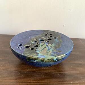 May include: A blue and green ceramic flower frog with a circular base and a pattern of holes for holding stems.