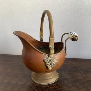 Pode incluir: Um balde de gelo de cobre e latão com uma alça de porcelana. O balde tem uma forma arredondada e uma base decorativa. A alça é presa ao lado do balde e tem uma maçaneta de porcelana.