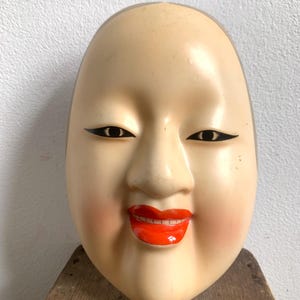 May include: A vintage Japanese Noh theatre mask with a serene expression. The mask has a pale complexion, black eyeliner, and bright red lips. The cheeks are blushed with a rosy hue, and it is set against a neutral background.