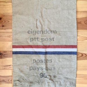 Può includere: Un sacco postale beige vintage con un design a strisce rosse, bianche e blu. Il sacco presenta il testo "eigendom ptt post" e "postes pays-bas 90". Ha anelli di metallo nella parte superiore per appenderlo.