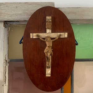 Puede incluir: Una placa ovalada de madera con un crucifijo dorado. El crucifijo presenta una figura de Jesucristo con una corona de espinas. La placa tiene una veta de madera marrón claro.