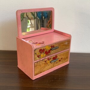 Pode incluir: Uma penteadeira de madeira rosa com um espelho e duas gavetas. As gavetas são decoradas com flores e folhas pintadas.