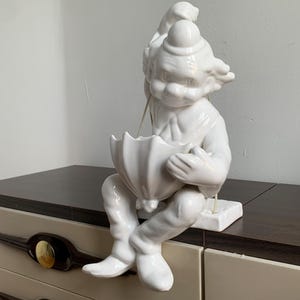 Puede incluir: Figura de cerámica blanca de un payaso sosteniendo un paraguas. El payaso lleva un sombrero y tiene una gran sonrisa. La figura está sentada sobre una superficie de madera.