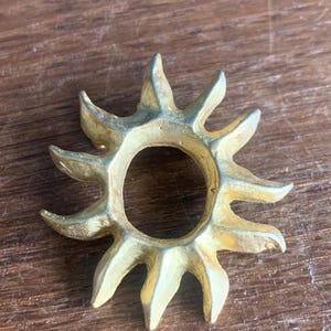 Puede incluir: Un anillo dorado en forma de sol con rayos puntiagudos. El anillo tiene una abertura circular en el centro y está sobre una superficie de madera. El diseño del sol es un elemento decorativo.