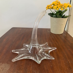 以下が含まれることがあります： 星型のベースと湾曲した螺旋状の茎を持つ透明なガラス製の装飾花瓶。花瓶は濃い木製の表面に置かれています。 黄色の花が入った小さな鉢植えも近くにあります。