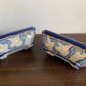 Puede incluir: Dos macetas de bonsái de cerámica azul y blanca con un diseño de palomas blancas volando en un cielo azul. Las macetas tienen una forma rectangular con bordes redondeados.