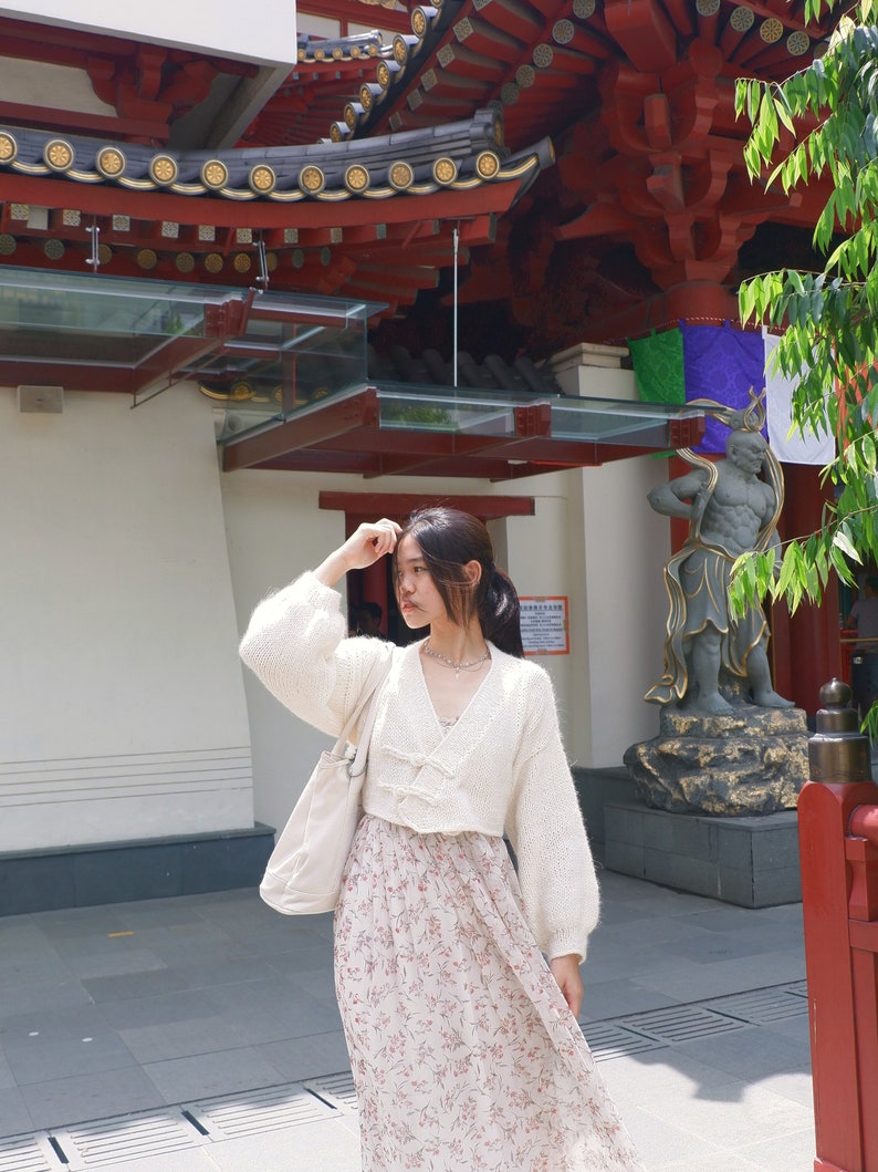 May include: A woman wearing a white knitted cardigan with a floral print skirt and a white tote bag. She is standing in front of a red and gold building with a statue.