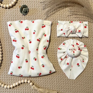 May include: A set of baby accessories, including a wrap, headband, and turban, all in white ribbed fabric. The items are adorned with a repeating pattern of red cherries and green leaves. The wrap has the text "Dear Me".