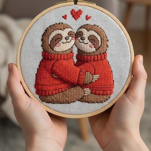 May include: Cross-stitch embroidery of two sloths in red sweaters, embracing. The sloths have brown fur, pink cheeks, and are kissing. Small red hearts float above them. The embroidery is set in a wooden hoop.