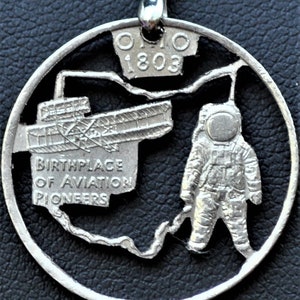 May include: A silver quarter coin necklace with a cutout design of an astronaut and a plane. The coin has the text "Ohio 1803" and "Birthplace of Aviation Pioneers" on it.