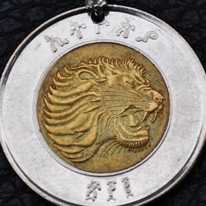 Puede incluir: Una moneda de un birr etíope de plata con una cabeza de león dorada en el anverso. La moneda tiene el texto "አንድ ብር" alrededor del borde de la moneda y "ብር" en la parte inferior.