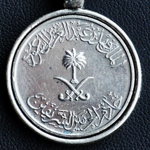 May include: A silver-colored coin pendant with Arabic script and a palm tree with crossed swords emblem. The coin is round with a detailed design and a small loop for a chain.
