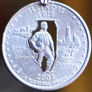 May include: A silver Illinois state quarter coin with a cutout of the state's outline. The coin features the text "ILLINOIS 1818" at the top, and "Land of Lincoln" and "21st State Century" on the sides.