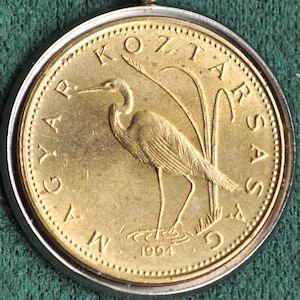 May include: A gold-colored coin with a silver rim featuring a bird standing in a pool of water. The coin has the text "MAGYAR KOZTARSASAG" and "1994" inscribed on it.