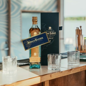 K&ouml;nnte beinhalten: Eine Flasche Johnnie Walker Blue Label Scotch Whisky mit einem goldenen Etikett und einem personalisierten Etikett mit der Aufschrift "Groom's Man James Brown" auf einer Holzoberfl&auml;che mit drei klaren Gl&auml;sern.