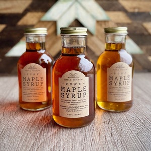 Puede incluir: Tres botellas de vidrio de jarabe de arce puro con tapas y etiquetas doradas. Las etiquetas dicen "Maple Hollow Pure Maple Syrup". El jarabe varía en color, desde ámbar claro hasta ámbar oscuro. El fondo presenta un diseño de estrella de madera.