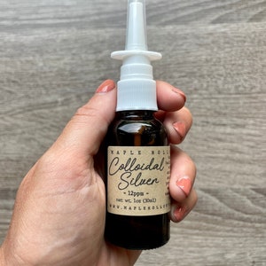 Könnte beinhalten: Braune Glasflasche mit weißem Nasenspray-Applikator. Das Etikett lautet "Colloidal Silver - 12ppm - net wt. 1oz (30ml)" mit dem Maple Hollow-Logo. Die Flasche wird in einer Hand vor einem Holzgrund gehalten.