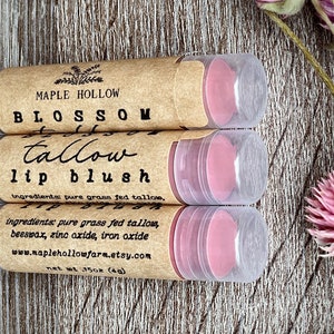May include: Three lip blush tubes with pink product inside, labeled "Maple Hollow Blossom Tallow Lip Blush." The tubes are in a brown paper casing with clear plastic lids. The ingredients are listed on the label. The net weight is 1.5 oz (4g).