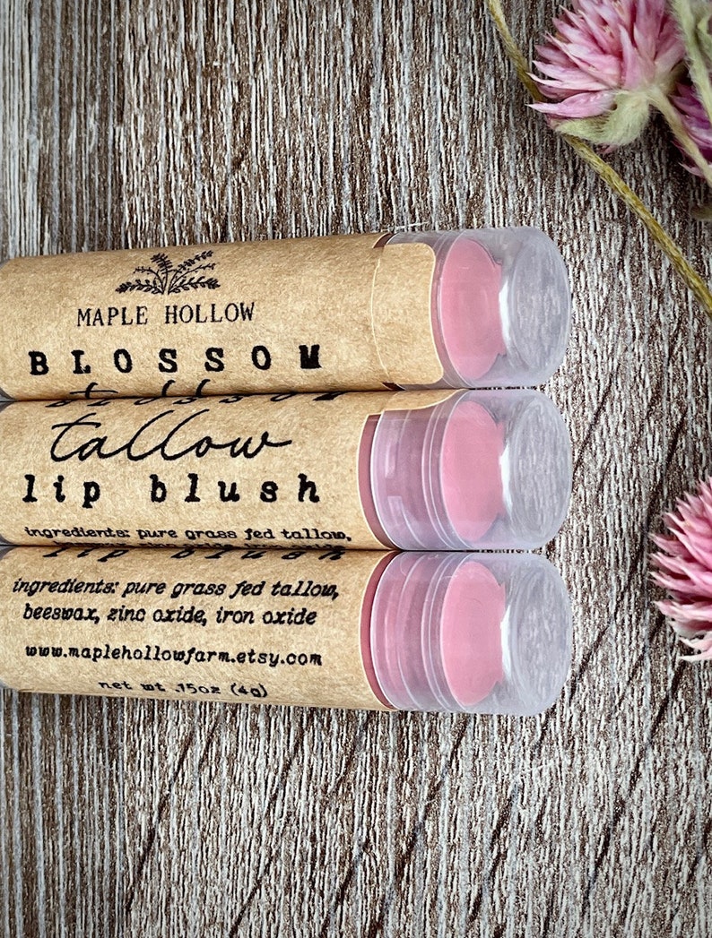 May include: Three lip blush tubes with pink product inside, labeled "Maple Hollow Blossom Tallow Lip Blush." The tubes are in a brown paper casing with clear plastic lids. The ingredients are listed on the label. The net weight is 1.5 oz (4g).