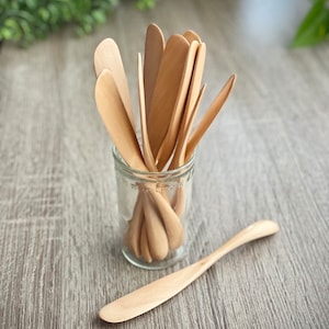 May include: A group of wooden butter knives in a glass jar, with one knife lying on a wooden surface.