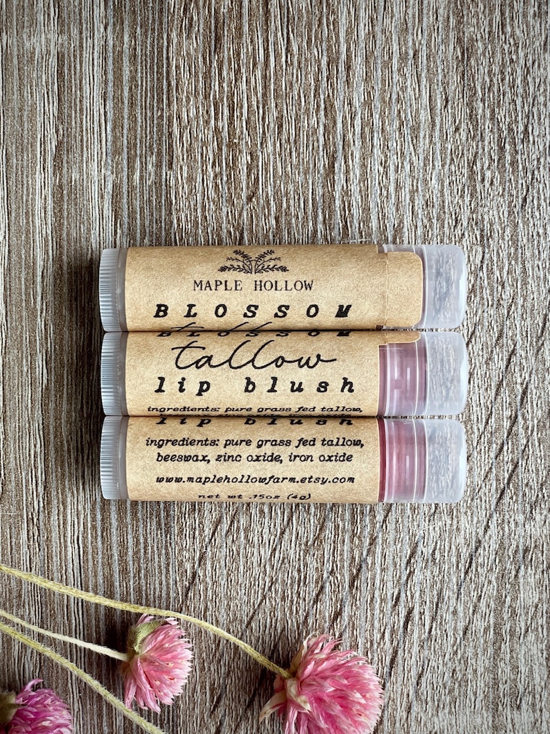 May include: Three lip balm tubes with brown labels and clear caps are stacked on a wooden surface. The labels read "MAPLE HOLLOW," "BLOSSOM," "tallow lip blush," and list ingredients. The net weight is 15oz (4g). Pink flowers are in the foreground.