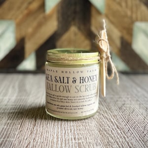 May include: A glass jar of "Sea Salt & Honey Tallow Scrub" with a wooden stirring stick tied to the lid with twine. The label includes the brand name "Maple Hollow Farm". The jar contains a light yellow substance and is 170 g.