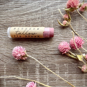 May include: A lip blush tube with the words "Blossom Tallow lip blush" printed on a brown label. The lip blush is a light pink color. Pink dried flowers are scattered around the lip blush on a wooden surface.
