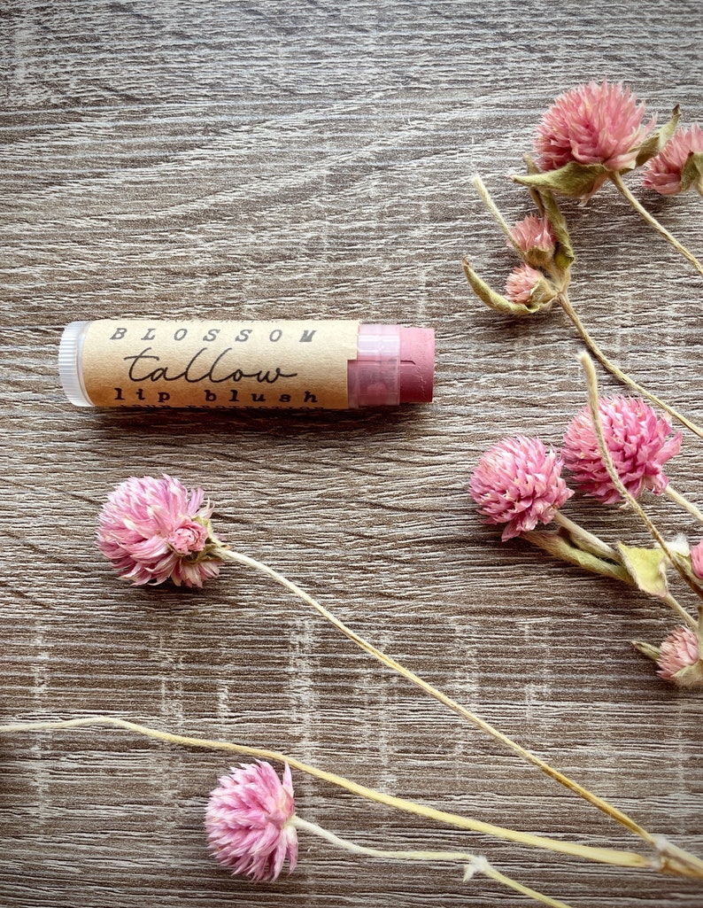 May include: A lip blush tube with the words "Blossom Tallow lip blush" printed on a brown label. The lip blush is a light pink color. Pink dried flowers are scattered around the lip blush on a wooden surface.
