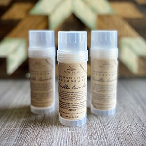 May include: Three clear plastic deodorant tubes with brown labels. The middle tube reads "Magnesium + Tallow Deodorant Vanilla Lavender" and lists ingredients. The tubes are on a light brown wooden surface.