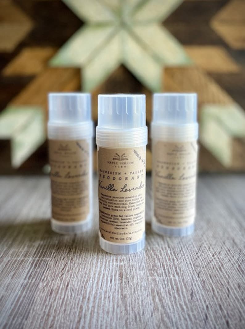 May include: Three clear plastic deodorant tubes with brown labels. The middle tube reads "Magnesium + Tallow Deodorant Vanilla Lavender" and lists ingredients. The tubes are on a light brown wooden surface.