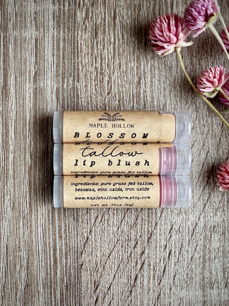 May include: Three lip balm tubes with brown labels and clear tubes are stacked on a wooden surface. The labels read "MAPLE HOLLOW BLOSSOM tallow lip blush." The ingredients are listed. Pink flowers are in the background.
