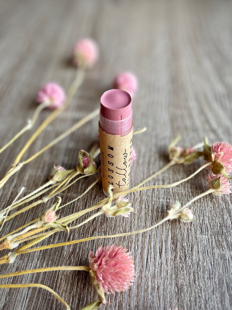 May include: A pink lip balm stick with a brown paper label that reads "Blossom Tallow" is surrounded by dried pink flowers and stems. The lip balm is on a wooden surface.