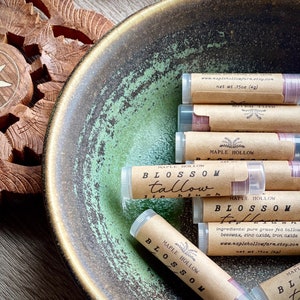 May include: A collection of lip blushes in clear tubes with brown paper labels, arranged in a green and brown ceramic bowl. The labels read "Blossom Tallow Lip Blush" and include the website address.