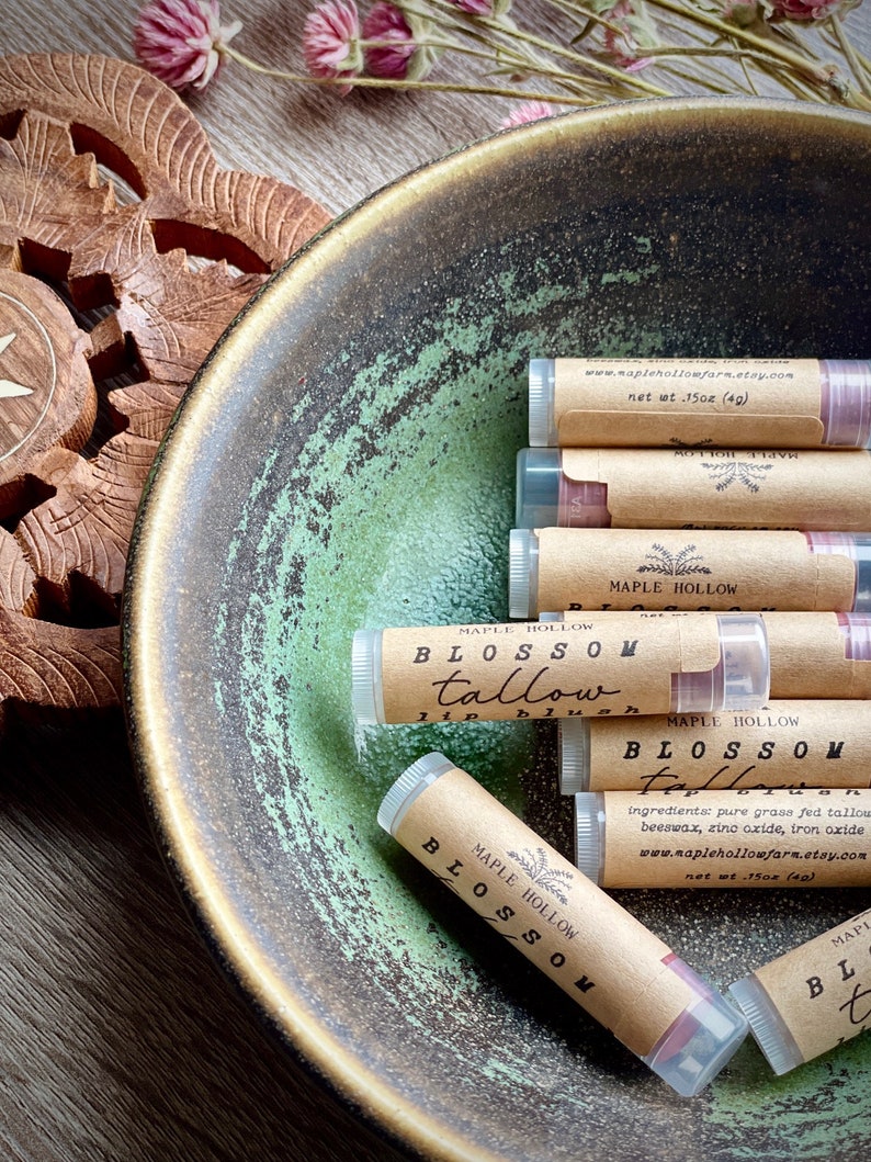 May include: A collection of lip blushes in clear tubes with brown paper labels, arranged in a green and brown ceramic bowl. The labels read "Blossom Tallow Lip Blush" and include the website address.
