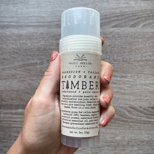 May include: A white deodorant stick with a brown label that reads "MAPLE HOLLOW FARM" and "MAGNESIUM + TALLOW DEODORANT TAMBER cedarwood + palo santo". The label also lists the ingredients and website address. The stick is 2 ounces (57g).