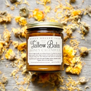 May include: A jar of Maple Hollow Farm Tallow Balm, Honey Calendula, with a gold lid and a label with product information. The jar is surrounded by dried yellow flowers on a wooden surface. The label includes the text: "www.maplehollowfarm.etsy.com net wt 5 oz. 142g."