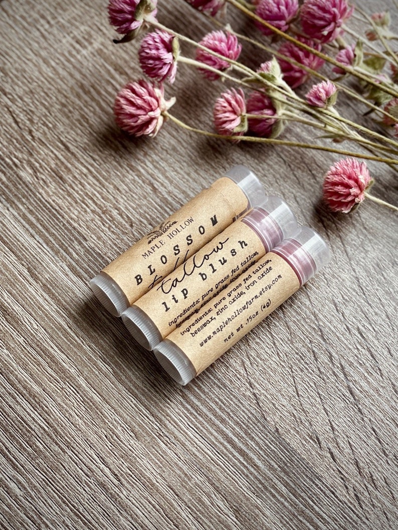 May include: Three lip blush tubes with brown labels and the text "BLOSSOM tallow lip blush" are arranged on a wooden surface. The labels also include the ingredients and website. Pink dried flowers are in the background.