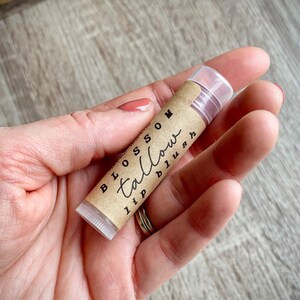 May include: A lip balm tube with the words "Blossom Tallow lip blush" printed on a brown label. The tube is held in a person's hand, with a clear cap and a light pink balm visible.
