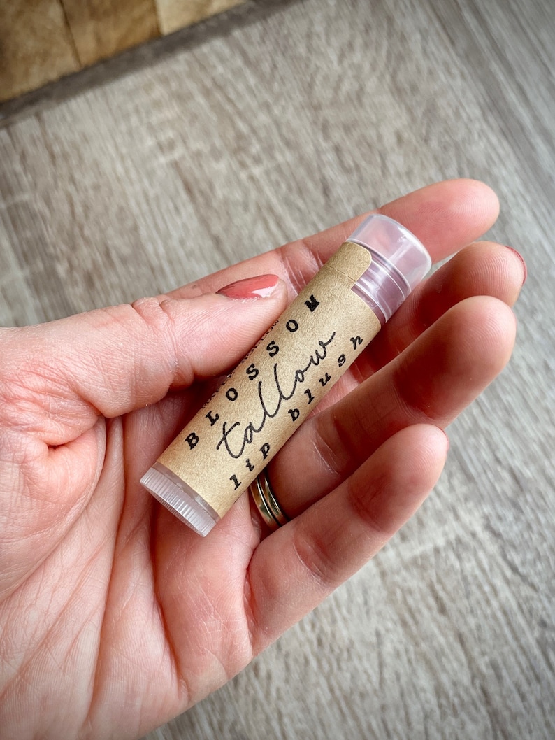 May include: A lip balm tube with the words "Blossom Tallow lip blush" printed on a brown label. The tube is held in a person's hand, with a clear cap and a light pink balm visible.