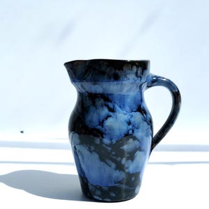 May include: A handmade ceramic jug with a dark blue and black marbled glaze. The jug has a curved handle and a slightly flared spout. The height is approximately 18 cm.