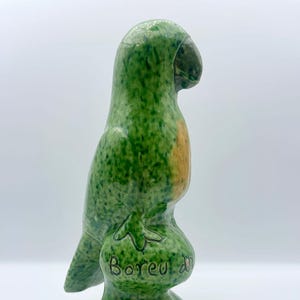 May include: A ceramic parrot figurine in shades of green with a touch of orange. The parrot is perched on a green base with the text "Boreu d" inscribed. The figurine has a glossy finish and a detailed beak.